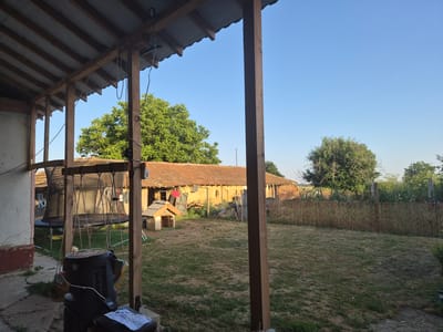 The image depicts a backyard scene in Kalipetrovo, Bulgaria, under a clear blue sky. In the foreground, a wooden porch with a corrugated metal roof is visible, supported by rough-hewn wooden posts. A black object, possibly a piece of discarded electronics or a large speaker, hangs from ropes attached to the porch structure, along with other miscellaneous items. Beyond the porch, a dry, grassy yard stretches out.

In the middle ground, a long, single-story building with a tiled roof and light-colored walls stands. Clothes are hanging on a line outside this building. To the left of this building, a small, wooden doghouse with a tiled roof is present. Further to the right, a fence made of bundled reeds or bamboo separates the yard from a more overgrown area.

The background features lush green trees and more of the tiled-roof building, suggesting a rural or semi-rural setting. The overall impression is one of a lived-in, somewhat cluttered backyard, possibly part of a residential property. The lighting suggests it is daytime, likely late afternoon or early evening given the warm tones. No people are visible in the image.