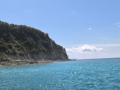 The image captures a scenic coastal view, likely near the city of Lermontovo, Russia. A steep, tree-covered cliff dominates the left side of the frame, meeting the turquoise waters of the sea. The cliff face appears rocky with visible strata. Along the base of the cliff, a narrow strip of rocky shore is visible, with a few small figures appearing to be standing or walking on it. The sea is a vibrant blue, with sunlight glinting off its surface, creating a shimmering effect. A few white clouds dot the clear blue sky. The overall impression is one of a tranquil natural landscape.