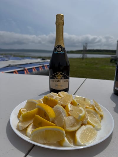 A bottle of Russian sparkling wine stands on a white table, behind a white plate filled with lemon wedges. The bottle is dark green glass with a gold foil top and a label that reads "РОССИЙСКОЕ". The plate of lemons is in the foreground, with the bottle slightly out of focus behind it. In the background, a body of water is visible, with a grassy shoreline and a cloudy sky. The scene appears to be outdoors, possibly near a waterfront. There are no people visible in the image. The location is identified as Murav'evo, Russia.