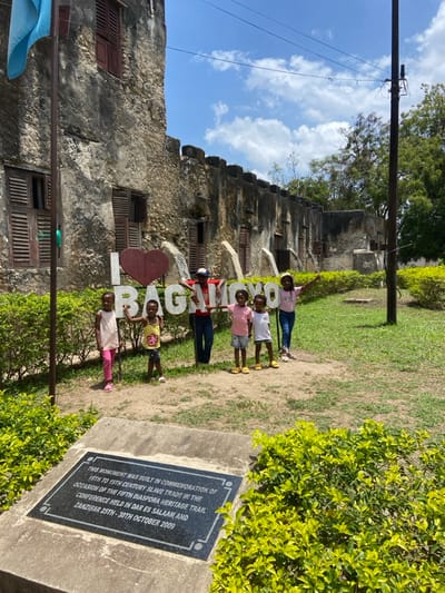 The image displays an outdoor scene in Bagamoyo, Tanzania, under a bright blue sky with scattered white clouds. Six children, ranging in age, are positioned in front of a large "I ❤️ BAGAMOYO" sign, composed of white letters and a dark red heart, arranged on a grassy and dirt patch. From left to right: a girl in a pink tank top and pants, a girl in a yellow tank top and patterned shorts, an older child in a red t-shirt and dark cap, a girl in a pink t-shirt and light shorts, a boy in a white t-shirt and light shorts, and a girl in a pink t-shirt and blue jeans. Several children have their hands on the letters of the sign, and all are facing the camera.

Behind the children and the sign, a weathered, multi-story stone building with a crenellated roofline and multiple arched windows fitted with dark louvered shutters stretches across the mid-ground. A metal flagpole, partially visible on the far left, holds a light blue flag. On the right, a dark utility pole with overhead wires extends upwards.

In the immediate foreground, a black rectangular plaque is set into a concrete base, stating: "THIS MONUMENT WAS BUILT IN COMMEMORATION OF 16TH TO 19TH CENTURY SLAVE TRADE IN THE OCCASION OF THE FIFTH DIASPORA HERITAGE TRAIL CONFERENCE HELD IN DAR ES SALAAM AND ZANZIBAR 25TH - 30TH OCTOBER 2009". The ground consists of green grass and patches of bare earth, bordered by manicured green bushes. Additional trees are visible in the distant background.