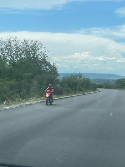 A motorcycle rider, wearing a red jacket and helmet, is riding on a grey asphalt road. The road curves around a patch of green bushes. The background shows a cloudy blue sky with distant hills. The location is the city of Makariopolsko, Bulgaria.