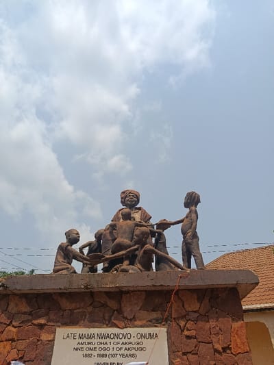 A reddish-brown weathered statue complex stands under a partially cloudy sky in Ndiaga-Akpugu, Nigeria. The central feature is a large statue depicting an older female figure, possibly a matriarch, seated at the top. She wears a headwrap and traditional attire, displaying a slight smile. Surrounding her are multiple smaller figures, appearing to be children or adolescents, engaged in a communal activity. One figure on the far left kneels, holding a long, thin object, while another on the far right stands, facing the central group, also interacting with a similar object. The figures appear to be collaborating with these objects, possibly representing agricultural implements or harvested goods.

The statue complex rests on a robust rectangular base constructed from rough-hewn reddish-brown stone blocks. Affixed to the front face of this base is a white rectangular plaque with black text, identifying the monument as "LATE MAMA NWAONOVO - ONUMA," born 1882 and deceased 1989, living 107 years. Her titles are listed as "AMURU OHA 1 OF AKPUGO" and "NNE OME OGO 1 OF AKPUGO." The plaque also states "UNVEILED BY" with further text not fully visible.

In the background, behind the statue, green foliage is visible on the left. On the right, part of a building with a reddish-brown tiled roof is present. Thin, dark utility wires stretch horizontally across the middle ground. The overall scene is illuminated by daylight.