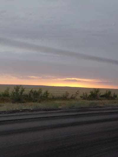 This is a landscape photograph captured at a low angle. The image depicts a road, a grassy field with scattered bushes, and a cloudy sky. The sun is setting, casting a warm glow on the horizon, creating a transition of colors from light blue to orange. The road, which occupies the right side of the frame, appears to be made of dark asphalt with visible tire tracks. To the left of the road is a field of dry grass, dotted with small, green bushes. The sky is filled with soft, gray clouds, with a bright yellow and orange streak of light near the horizon, indicating the setting sun. The overall mood of the image is serene and peaceful, evoking the calm of a rural sunset. There is no visible text in the image. The location context suggests this scene is from Қарасу ауылдық округі, Kazakhstan.
