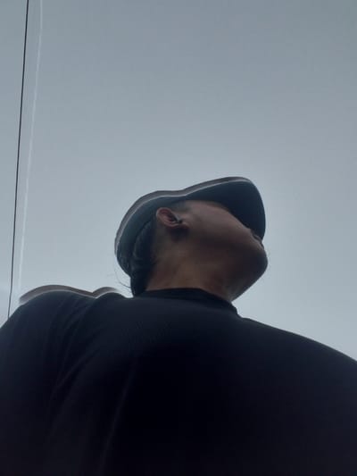 The image shows a person from a low-angle perspective, looking upwards against a light gray sky. The person is wearing a black t-shirt and a dark baseball cap. A thin black line, possibly a wire or cable, runs vertically on the left side of the image. There is a faint vertical streak of light to the right of the black line, which may be a reflection or a contrail. The person has dark hair visible under the cap and is wearing an earbud. The overall scene is outdoors, with the sky occupying the majority of the frame. The context of Kubang Jaya, Indonesia is not evident from the visual information provided in the image.