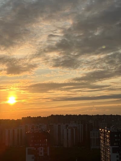 The image shows a city skyline at sunset in Feklistovo, Russia. The sky is filled with dramatic clouds, illuminated by the bright orange sun on the left side of the frame. Below the sky, a dense collection of buildings, likely apartment blocks, stretches across the horizon. The buildings vary in height and architectural style, with many featuring multiple windows. Some buildings are silhouetted against the bright sky, while others are partially illuminated by the setting sun, creating a play of light and shadow. The overall impression is of an urban landscape bathed in the warm, atmospheric light of dusk.