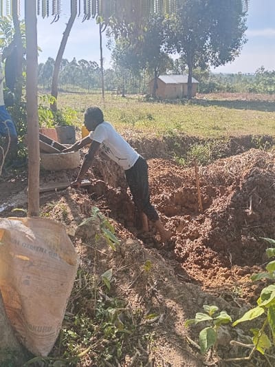 A person is seen outdoors near an earthen structure. They wear a white t-shirt and dark pants and appear to be working or interacting with the ground. A bag sits nearby, and vegetation is visible in the surrounding area. In the background, there's a structure, possibly a building, framed by trees and a bright sky. The photo is taken in a bright, sunny condition.