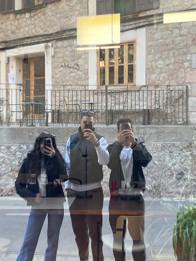 A reflection of three individuals is captured in a glass window in Fornalutx, Spain, with the exterior of a stone building serving as the background.
The three individuals, appearing to be two males and one female, are positioned frontally, holding smartphones with both hands, pointed outwards as if taking a photo or video.
1. **Left individual:** A female with long, dark, wavy hair, wearing a dark jacket over a white top and blue jeans. Dark sunglasses are perched on her head.
2. **Center individual:** A male with dark, short-to-medium length hair, wearing a dark olive green vest over a white long-sleeved shirt and dark brown pants. Dark sunglasses are perched on his head, and a ring is visible on his left ring finger.
3. **Right individual:** A male with dark, short hair (partially obscured by his phone), wearing a dark jacket over a white top and light brown/tan pants.
Through the reflective glass, the background reveals the facade of a traditional stone building. A wooden-framed entrance door is visible on the left, with faint lettering ("Cantina" or similar) above it. A multi-paned wooden window is present on the right, topped by closed brown shutters. Below these, a raised outdoor terrace or balcony features a black metal railing and several dark metal tables and chairs, suggesting a cafe or restaurant. A paved surface is faintly visible at the bottom of the background. Reflections of rectangular yellow light panels from inside the establishment are visible in the upper portion of the glass. A potted green plant is partially visible in the bottom right foreground.