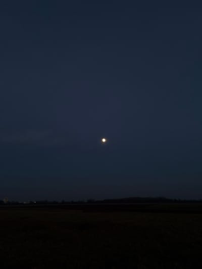 A dark, dusky sky dominates the view. A small, bright moon is positioned near the center. The rightmost part of the image is very dark, seemingly a field or landmass obscured by shadows. A faint, lighter line separates the sky from this dark area, possibly indicating the horizon or a tree line. Overall, the image evokes a sense of nighttime serenity.
