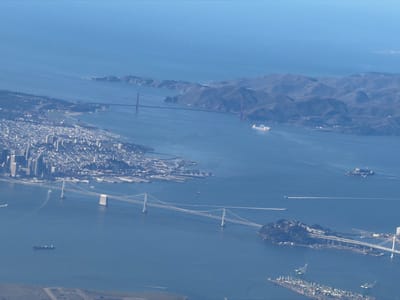 This aerial photograph displays a high-altitude view of the San Francisco Bay Area, United States, under clear daylight conditions. The scene encompasses a vast body of water, identified as the San Francisco Bay and a segment of the Pacific Ocean, bordered by multiple landmasses.
In the upper-center, the Golden Gate Bridge, a red suspension bridge, spans the entrance to the Pacific Ocean. To the left, the urban density of San Francisco is visible on a peninsula, characterized by numerous buildings forming a distinct skyline. The San Francisco-Oakland Bay Bridge, a multi-segment white suspension structure, extends from San Francisco, crosses the tree-covered Yerba Buena Island, and continues eastward towards Oakland. Alcatraz Island, featuring identifiable structures, is positioned in the mid-right section of the bay.
The foreground of the image, located within the city of Oakland, depicts active industrial port infrastructure. This includes multiple green gantry cranes positioned along piers, adjacent to visible stacks of cargo.
The water surface of the bay contains several vessels: a large white cargo ship moving near the Golden Gate Bridge, a gray naval-looking vessel in the mid-right, and various smaller boats, some generating visible white wakes. No individual people are discernible due to the altitude.