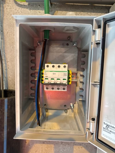 A white electrical enclosure box is open, revealing a circuit breaker panel. The enclosure is mounted on a textured concrete wall, with some conduit and wiring visible above and to the left of the box. Inside the enclosure, a DIN rail system is populated with several Schneider Electric circuit breakers. A bundle of three electrical wires, colored blue, brown, and grey, enters the enclosure from the top and appears to be connected to the circuit breakers. The enclosure itself has a grey inner panel where the circuit breakers are mounted. The lid of the enclosure is open and shows a label with "Schneider Electric" and a product code. The scene appears to be an outdoor or utility area, possibly near industrial facilities, given the concrete wall and conduit. The lighting suggests it is daytime.