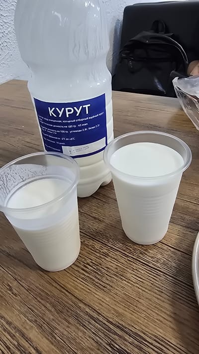 A close-up, eye-level shot shows a white plastic bottle of fermented baked milk and two clear plastic cups filled with the same product, arranged on a wooden table. The bottle has a blue label with white Cyrillic text, which appears to be product information. The cups are positioned in front of the bottle, with one slightly to the left and the other slightly to the right. The background is blurred, but appears to be an indoor setting with a dark chair and a black backpack. The lighting is natural and diffused, creating soft shadows. The image composition focuses on the dairy products, with the bottle and cups dominating the foreground. The overall scene suggests a casual consumption of the beverage, perhaps at home or in a cafeteria. The location is specified as Fryazevo, Russia.