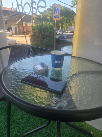 A smartphone and a pack of cigarettes with a lighter sit on a glass-topped table outside a cafe with the word "dose" visible in large lettering on a window. The cafe is located in Khasavyurt, Russia. The table is set on a patch of artificial grass, and in the background, there is a street with cars and buildings. The scene is bathed in sunlight, casting reflections on the glass table.