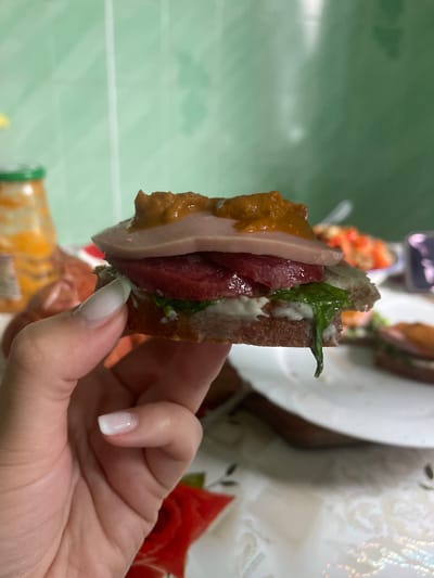 A close-up shot shows a hand with well-manicured nails holding a small, layered sandwich. The sandwich consists of a dark bread base, topped with what appears to be white sauce, green leafy vegetables, a slice of red meat, a slice of pale pink meat, and a dollop of a dark, chunky sauce on top. In the blurred background, a glass jar with an orange substance, a plate with food, and a green tiled wall are visible. The image appears to be taken indoors, likely in a kitchen or dining area. The location within the city of Красное, Russia is not discernible from the visual information provided in the image.