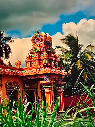 An ornate, brightly colored Hindu temple, likely in Natal, Brazil, is the central subject. The temple features a multi-tiered, intricately decorated tower (Vimana) with statues and carvings. The building's facade is also adorned with vibrant patterns and colors, including red, orange, yellow, and green. Palm trees frame the temple on the left and right, with lush green foliage and tall grass in the foreground partially obscuring the lower portion of the scene. The sky above is a mix of bright blue and dramatic clouds, suggesting a dynamic weather condition. A few figures are visible near the entrance of the temple, though their features are indistinct.