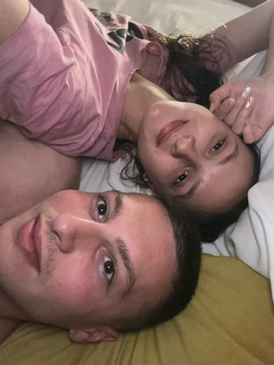 A selfie of two people lying in bed. The man is at the top of the frame, with short hair, and fair skin. The woman is positioned at the bottom, with dark curly hair and fair skin. Both are looking into the camera. The man is shirtless, while the woman is wearing a pink t-shirt. They are lying on white sheets with a yellow pillow visible behind the man's head. The location is Vitry-sur-Seine, France, which is not discernible from the image.