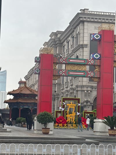 The image depicts a street scene in Shenhe District, China, likely a commercial or tourist area. A large traditional Chinese archway, adorned with red, gold, and decorative blue and green accents, dominates the foreground. To the left of the archway stands a multi-story building with grey stone and elaborate architectural details. To the right of the archway, a planter with a small tree sits on a raised concrete area. On the far right is a low white fence. Underneath the archway, there are decorative floral arrangements and what appears to be a large sign with yellow Chinese characters. People are walking around and sitting on the ground. Further down the street beneath the archway is a traditional Chinese building. In the background, there are modern high-rise buildings. The sky is overcast.