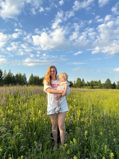 A woman is standing in a meadow filled with tall green grass and various wildflowers, including purple lupine and yellow flowers. She is holding a young child in her arms. The woman has long, wavy blonde hair and is wearing a white t-shirt and white shorts. The child has blonde hair and is wearing a light-colored romper and has a pacifier in their mouth. In the background, there is a line of trees under a bright blue sky with scattered white clouds. The image was taken in the village of Koloksha, Russia.