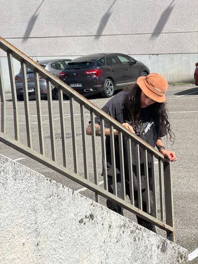 The image shows a person wearing a brown corduroy bucket hat and a black t-shirt with white graphic print. The person has long, dark, curly hair and visible tattoos on their arm. They are leaning over a metal grate structure, which appears to be a fire escape or a set of stairs. The person is holding a small orange object in their right hand.
The scene is set outdoors, with a paved surface that resembles a parking lot visible in the background. Two dark-colored cars are parked in the background, with one car's license plate visible reading "ET-006-NN". To the left of the person, there is a textured wall, possibly of a building. The lighting suggests it is daytime, with strong shadows cast on the ground.
The person's posture and gaze suggest they are engaged in an action or observing something below. The overall mood is casual and perhaps a bit pensive.
Visible text includes the word "Makers" on the person's hat. The location context provided is Caluire-et-Cuire, France.