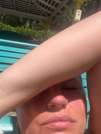 The image presents a close-up, high-angle view of an adult individual in Olde Yard Village, British Virgin Islands. The person's right arm, displaying a light skin tone with fine hair, extends horizontally across the upper face, obscuring the eyes and forehead, acting as a sun shield. The visible lower half of the face shows sun-reddened skin, particularly on the nose, which features a gold-colored hoop nose ring on the right nostril. The face appears moist. The lips are closed. In the background, a light blue slatted bench or lounger is partially visible. Beyond this, a yellow support beam and a white slatted roof structure, indicative of a gazebo or pergola, are present. A beige wall-mounted dispenser with a dark circular opening is affixed to the yellow beam. Green foliage, illuminated by bright sunlight, is visible further in the background.