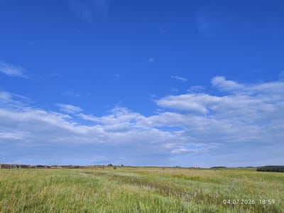The image depicts a rural landscape under a bright blue sky with scattered white clouds. In the foreground, a grassy field stretches out, with a path or less dense vegetation running through it. In the middle ground, a weathered wooden fence, possibly part of a farm or enclosure, runs horizontally across the frame, with various wooden posts and structures visible. Beyond the fence, the land appears to undulate and is dotted with sparse vegetation and some distant trees. The horizon is marked by rolling hills and a line of trees on the far right. A timestamp in the bottom right corner reads "04.07.2026 18:59". The scene suggests a peaceful, natural environment in the countryside of Konovalovo, Russia. No people are present in the image.