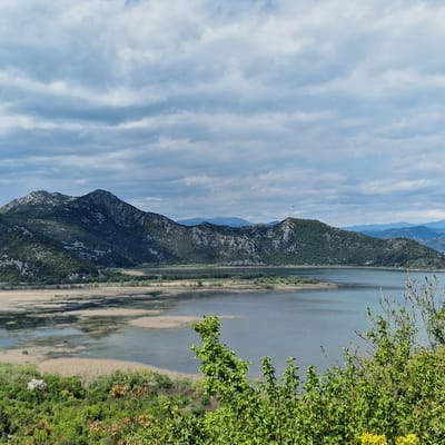 The image shows a landscape in Godinje, Montenegro. A lake stretches along the right side of the frame, next to a grassy and sandy shoreline. Green foliage grows densely on the right edge of the image. A mountain range runs parallel to the lake on the left, with a sky of pale blue and streaky white clouds in the background.