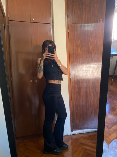 The image is a rotated mirror selfie taken indoors. A person, appearing to be lying horizontally due to the image's 90-degree rotation, is the central subject. The person has dark hair and is wearing a black cropped top, black pants, and black athletic shoes. Tattoos are visible on their arms and possibly along their rib cage. They wear a watch on their left wrist and rings on their fingers. Their face is obscured by a dark-colored smartphone, which they are holding up to take the picture.
The background, as seen in the mirror's reflection, consists mainly of dark, polished wooden panels, likely part of a wardrobe or cabinet, featuring visible wood grain and small metal hinges. Below these panels is a light-colored wall or baseboard. To the right of the person, a patterned wooden parquet floor is visible. A bright light source, possibly a window, creates a glare in the upper portion of the mirror's reflection. The mirror itself has a thick black frame. No visible text is present. The setting is indoors, likely daytime, in Llavallol, Argentina.