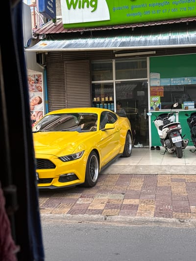 A bright yellow Ford Mustang is parked on a sidewalk in front of a shop in Khan Boeng Keng Kang, Cambodia. The shop has a green awning with white lettering that says "Wing." To the right of the Mustang, a white scooter is parked. The sidewalk is made of patterned tiles. A person is visible through the shop window in the background. The car's reflection is visible in the shop window.