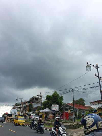 The image depicts an urban street scene in Palayan, Philippines, under a heavily overcast sky. The upper two-thirds of the frame are dominated by dense, dark grey clouds, indicating overcast weather.
In the foreground, the back of a person wearing a blue and white motorcycle helmet is visible on the far right, facing away from the viewer. The asphalt road occupies the lower left and central portions of the image, marked by a solid yellow line on the left edge. Traffic is present, with multiple motorcycles traveling away from the viewer. A white scooter with a single rider wearing a white helmet is prominent in the lower right, followed by a dark scooter carrying two individuals (driver and passenger). Further back, another dark scooter with two riders is visible. A yellow passenger van is seen traveling in the opposite direction on the left side of the road, with a dark sedan partially visible behind it.
The roadside on the right features a concrete barrier separating the road from a raised area where commercial structures are situated. These include a white tent, an adjacent structure with a red roof, and a visible sign bearing the text "BUKIT CINTA" (partially obscured), mounted on a pole. Numerous utility poles with a complex network of overhead black wires crisscross the scene. A decorative streetlight, featuring a green-tinted lantern with gold-colored accents, stands on the far right. Along the left side of the street, various buildings are visible, including a multi-story, unfinished concrete structure with exposed rebar, and other buildings with red roofs, interspersed with trees and green vegetation. The overall environment suggests a bustling commercial area.