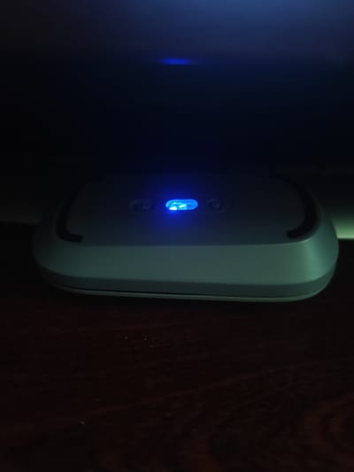 The image shows a close-up view of a light gray electronic device resting on a dark wooden surface. The device is oblong with rounded edges and features two circular buttons on its top surface. A bright blue indicator light is illuminated on the right side of the device, near one of the buttons. The background is dark and out of focus, with some faint blue light reflections visible, suggesting the device might be emitting light or reflecting ambient light from a screen or other light source. The overall mood is dim and somewhat mysterious due to the low lighting. There is no visible text in the image. The setting appears to be indoors, possibly a home or office environment. No people are visible, and the focus is entirely on the electronic object. The image does not provide specific clues about the time of day or weather.