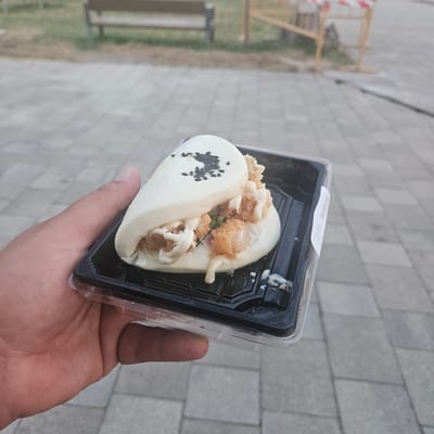 A hand is holding a black plastic container with a single piece of bao bun sandwich filled with fried shrimp and a white creamy sauce. The bao bun is soft and white, folded in half, with black sesame seeds sprinkled on top. The fried shrimp pieces are golden brown and visible within the bun, along with some green leafy vegetables. The creamy sauce is drizzled over the filling and drips down the side. The container has a white sticker on the side with some printed text. The background appears to be a paved area with grey square tiles, suggesting an outdoor setting. In the blurred background, there is a wooden bench and a yellow barrier with red and white tape. The image was taken in El Perelló, Spain, likely in a public area such as a park or promenade.