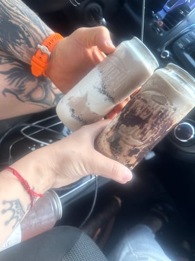 Two hands are holding two clear glass jars filled with drinks. The hand on the left has tattoos and is wearing an orange watch. The hand on the right has a red string bracelet with a crown charm. The jar in the foreground contains a chocolate-colored drink with swirls of syrup. The jar in the background contains a lighter-colored drink with what appears to be chia seeds. The background shows the interior of a car, with the dashboard and steering wheel visible. The scene is likely in Glenview, Illinois, United States of America.