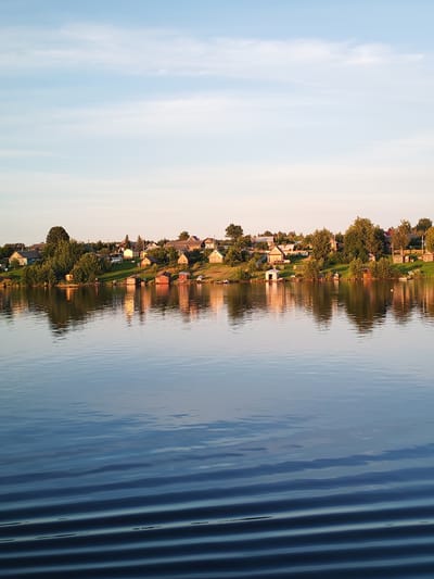 The image depicts a serene lakeside scene in the village of Bol'shaya Stepanovskaya, Russia. The foreground is dominated by the calm, reflective surface of a lake, with subtle ripples distorting the mirrored image of the opposite bank. The far bank of the lake is lined with a collection of small, rustic houses and sheds, nestled amongst lush green trees and vegetation. The houses appear to be primarily single-story structures, with pitched roofs, some painted in light colors and others made of wood. Several small boats are visible near the shore, suggesting a fishing or leisure community. The sky above is a soft gradient of pale blue and pink, indicative of either early morning or late evening light, with wispy clouds scattered across it. The overall impression is one of peaceful, rural tranquility.