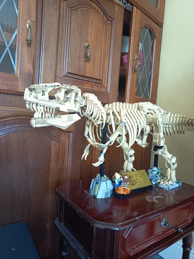 A LEGO Tyrannosaurus Rex skeleton model is displayed on a dark wooden side table in front of a large wooden cabinet. The cabinet has two doors with diamond-paned glass panels and decorative metal handles. The T-Rex model is beige and rests on black stands. In front of the T-Rex, on the table, are two LEGO minifigures, one of a woman and one of a man. Beside them is a gold plaque with "JURASSIC PARK" and information about the Tyrannosaurus Rex. The table has a glossy, dark brown finish and shows some signs of wear, including a light circular mark and dust. The wall behind the cabinet is a pale yellow. The scene is indoors, likely in a home setting. This location is in Millstreet, Ireland.