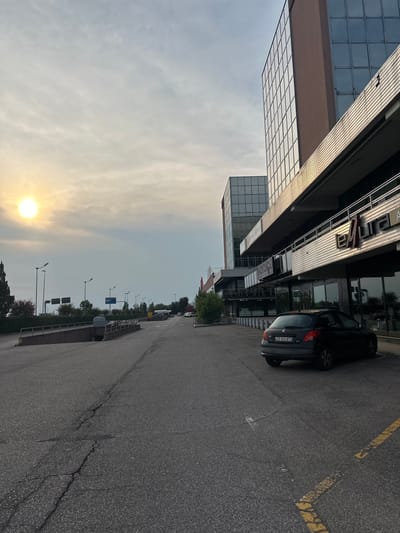 This media file shows a street scene in San Martino Buon Albergo, Italy. The image is oriented vertically, with the sky taking up a significant portion of the frame. The sun is visible in the lower left of the image, indicating it is either sunrise or sunset. The sky is hazy, suggesting overcast conditions or the presence of pollution.
On the right side of the frame, a dark-colored car is parked on the side of an asphalt road. The car has a license plate that reads "DS 622AT". Yellow lines are painted on the road, demarcating parking spaces or lanes. Further down the road, several streetlights are visible, as well as some trees and buildings.
To the left of the road, a modern building with a glass facade and metallic accents is prominent. The building appears to be several stories high. Part of the building has a sign with some visible text, though it is difficult to read clearly.
The scene appears to be a typical urban or suburban environment. The overall mood is neutral, with no discernible emotional cues. The time of day is either early morning or late evening due to the sun's position.