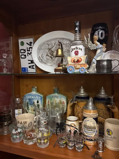 A shelf displays a collection of various objects. A Jim Beam bottle sits next to a porcelain figure and a "Fabulous 40" celebratory piece. Below, a plate with a deer motif rests above a license plate reading "AW 354". Adjacent, German beer steins and shot glasses, some labeled "Germany" and "1922", are arranged. Other glasses with labels like "Kansas" and "VATGO" are also present. The shelf appears to be within a wooden cabinet with a glass door.