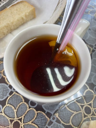 A close-up overhead shot shows a white mug filled with dark liquid, likely tea, with a slice of lemon floating in it. A silver spoon with a pink straw is partially submerged in the mug. Next to the mug, a slice of white bread sits on a white napkin, which is itself in a basket. The mug rests on a dark placemat with a decorative gold and grey floral pattern. The overall scene suggests a casual meal or break, possibly in a home or cafe setting. The lighting appears bright, indicating daytime. There is no visible text in the image.