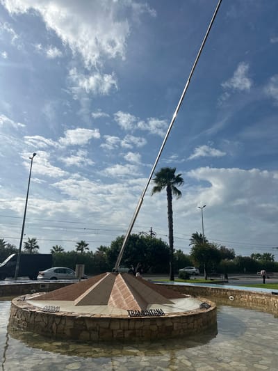 The image depicts "El Rellotge de Sol 'Tramuntana'" (The Tramuntana Sundial) located at Plaça de Catalunya, Pineda de Mar, Spain.

The central subject is a large circular water fountain containing a multi-tiered, pyramid-shaped monument. The fountain features a rough-hewn stone base and clear, shallow water reflecting the sky. The central monument is constructed from light-toned, possibly tiled, material. A long, slender, reflective metal gnomon extends diagonally upwards from the monument towards the upper right of the frame. The word "TRAMUNTANA" is displayed in metal lettering on the base of the central structure within the fountain.

In the background, a street is visible, populated by several vehicles including a dark van, a silver sedan, and a white SUV. Overhead electrical or tram lines span the mid-ground. Multiple tall palm trees and various green bushes line the street. A tall streetlight is present on the left, and another is visible on the right. A unique detail is a metallic bicycle sculpture mounted high on a pole, visible near the canopy of a prominent tall palm tree in the center-right background. On the far right, a single individual is standing by the roadside. The sky is partly cloudy, featuring a bright blue expanse with scattered white cumulus clouds. Strong sunlight is evident, creating reflections on the gnomon and the water surface.