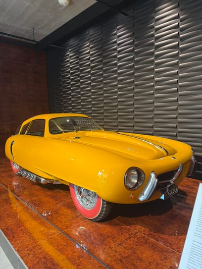 An extreme wide shot captures a bright yellow, vintage sports car on display in a museum. The car is positioned diagonally on a polished, wood-grained floor that reflects its vibrant color. The background features a textured, dark gray wall composed of undulating, horizontal elements that create a ripple effect. The car's design is sleek and aerodynamic, with prominent chrome accents, including a grille, bumper, and side exhaust pipes. It has a distinctive glass canopy and red-rimmed tires with spoked wheels. A placard with text is visible on the right side of the frame, partially obscuring the car's front passenger-side wheel. The lighting in the scene is dramatic, with spotlights highlighting the car and casting shadows. The overall impression is one of a meticulously presented exhibit in a modern museum setting. The car is likely on display at the Louwman Museum in Wassenaar, Netherlands, a renowned automobile museum.