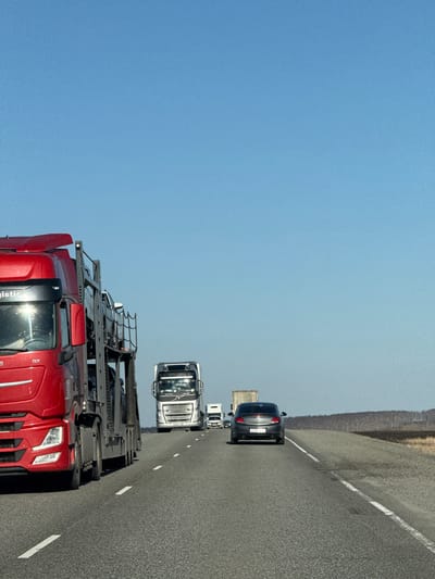 The image, when correctly oriented, depicts a clear, bright daytime scene on a multi-lane asphalt highway. The sky is a uniform, light blue, indicating fair weather.

In the foreground, a prominent red semi-truck tractor unit occupies the left lane. It features a distinctive front grille and headlights, characteristic of Volvo trucks, with a "GX" emblem visible near the driver's door and partially visible "Logistic" text on its side. A complex, multi-tiered rack structure is mounted above its cab.

Further down the road in the right lane, a silver or white semi-truck tractor unit, also appearing to be a Volvo, follows. Behind it, a dark-colored sedan, possibly an Opel Insignia, is seen with a partial view of its rear license plate, which includes characters like "C141", consistent with a Russian plate given the location context. A smaller white vehicle and a brown/tan semi-trailer are visible further in the distance.

The road itself is asphalt with clear white dashed lane markings. To the right of the highway, there is a gravel or dirt shoulder, and beyond that, a low horizon line with sparse, dark vegetation or trees under the wide-open sky.

The scene captures vehicles in traffic, either moving slowly or queueing. Sunlight glints off the surfaces of the vehicles, confirming it is daytime. There are no identifiable people visible, and the overall mood is neutral, showing an ordinary highway environment in Ol'gino, Russia.