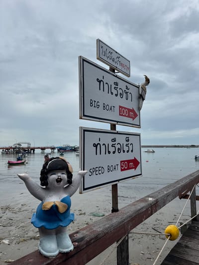 The image shows a pier in Ban Phe, Thailand, on an overcast day. In the foreground, a ceramic figurine of a woman with black curly hair, wearing a blue dress and white boots, stands on a wooden railing. To the right of the figurine are two white directional signs with black lettering. The top sign, angled slightly, has Thai script and reads "BIG BOAT 100 m." with a red arrow. The bottom sign reads "SPEED BOAT 50 m." with a red arrow. A wooden bird sculpture is perched on the pole holding the signs. The pier extends into a body of water with several boats docked and anchored, including fishing boats and a larger passenger boat. The water is calm, and the shoreline is visible with sand and debris. A yellow buoy is attached to ropes on the wooden railing in the lower right quadrant of the image.