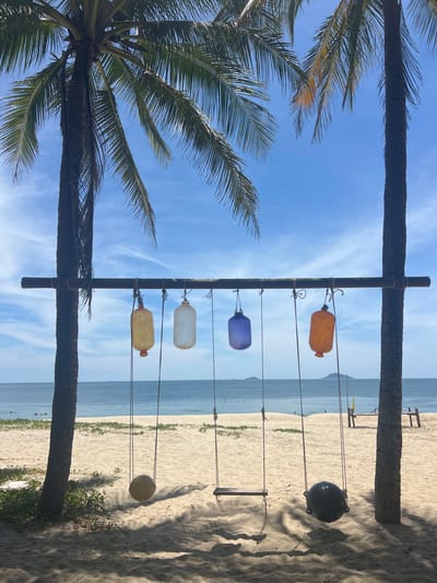 This image depicts a tropical beach scene, likely in Hoi An, Vietnam, characterized by palm trees, a sandy beach, and the ocean. In the foreground, a rustic wooden beam is suspended between two palm trees. Hanging from this beam are five decorative, translucent lanterns in various colors: yellow, clear, blue, and orange. Beneath the lanterns, three swings are suspended by ropes from the same beam. One swing seat is visible, while another appears to be a black, rounded object.

The sandy beach stretches towards the calm, blue ocean, where a few people are visible swimming in the distance. Two small islands or landmasses are visible on the horizon. The sky is a clear blue with some wispy white clouds. Shadows of palm fronds fall across the sand, indicating dappled sunlight. The overall impression is one of a peaceful and idyllic beach setting.