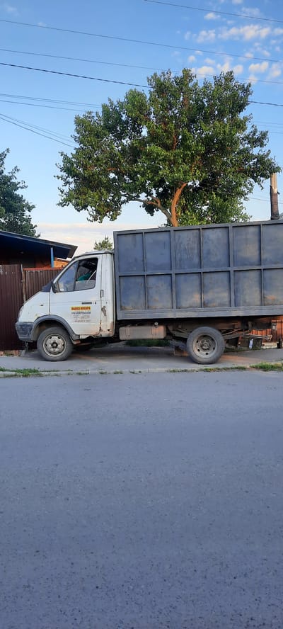 A side view of a white truck with a dark grey, open-top cargo bed is parked on a paved road next to a sidewalk. The truck has a logo on its side with text that appears to be in Cyrillic script, including "ВЫВОЗ МУСОРА" (Waste Removal) and "ГРУЗОПЕРЕВОЗКИ" (Freight Transport). The truck's cab is white, and a person's head and shoulders are visible inside, suggesting they are the driver or a passenger. Behind the truck, a large green tree with full foliage stands prominently against a blue sky with scattered clouds. To the left of the truck, a brown wooden fence and a portion of a building are visible. The environment suggests a day with clear weather.