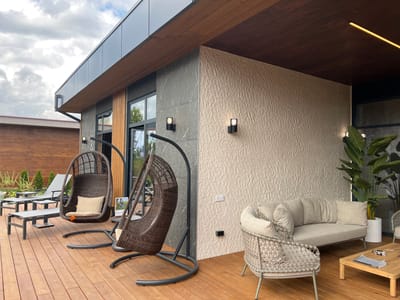 The image depicts an outdoor patio area in Yakunino, Russia, featuring modern outdoor furniture. The patio is constructed from wooden planks. In the foreground, there are two dark brown, hanging rattan chairs, each with a beige cushion. To the left, two gray lounge chairs with adjustable backs are placed on the deck. Further back, a light gray loveseat with multiple cushions is situated against a textured cream-colored wall. In front of the loveseat, a low, light brown wooden coffee table holds a magazine and a few small items. To the right of the loveseat, a large potted plant with broad green leaves is visible. The exterior of the building has a dark gray stone facade and a wooden soffit under a dark gray overhang. Two modern black outdoor sconces are mounted on the facade, one on the gray stone and another on the cream-colored wall. A thin, illuminated yellow strip light is installed along the wooden ceiling. The sky above is partly cloudy.