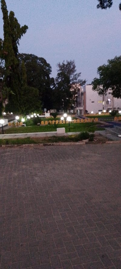 The image captures an outdoor scene at dusk or dawn in Tudor ward, Kenya. In the foreground, a dark-colored, rectangular-patterned paved surface is visible. This surface transitions to a low, light-colored concrete curb, separating it from a landscaped area. The midground features a garden with green grass and numerous spherical, illuminated outdoor lights mounted on short poles. Behind these lights, a curved arrangement of yellow, arch-shaped elements is present. Dense, dark-foliage trees occupy the left and central portions of the midground and background. On the right side of the background, a multi-story, light-colored building with several illuminated windows is visible. The sky above is a light blue-grey. No individuals or discernible activities are present.