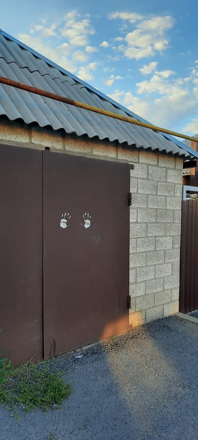 The image shows a close-up view of a garage door and a section of a brick wall, with a corrugated metal roof visible above. The garage door is a dark brown color and appears to be made of metal. On the left half of the garage door, there are two white handprints. To the right of the garage door, a brick wall is constructed from light-colored, rectangular bricks. A rusty pipe runs horizontally across the top of the roof. The sky above is blue with scattered white clouds. A small patch of green vegetation is growing in the bottom left corner of the image, next to the garage door. The ground in front of the garage appears to be covered in gravel. The scene is outdoors and appears to be in a residential or industrial area. The image does not provide enough context to determine the specific location within Shakhty City District, Russia, other than it is an outdoor scene.