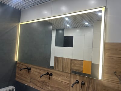 The image shows a modern bathroom with a large, backlit mirror above a wooden vanity. The vanity has two black faucets installed. The walls are a combination of grey tiles and a wood-grain patterned panel. The ceiling is a white grid. The mirror reflects the opposite wall, which features a geometric design of white, grey, and orange tiles, and a black square. The overall scene is clean and contemporary. The location is a bathroom in the city of Druzhny, Russia. There are no people visible in the image.
