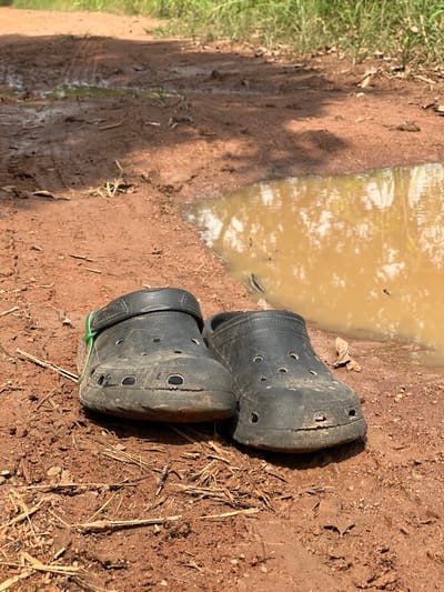 A pair of black, well-worn, and dirt-covered Crocs-style clogs rests on a reddish-brown unpaved dirt path in Logba Adzakoe, Ghana. The clogs show signs of heavy use, including surface abrasions, small cracks, and accumulated dirt. A green and red striped band is wrapped around the adjustable strap of the left clog. Adjacent to the clogs, on the right side of the frame, is a large muddy water puddle, reflecting the sky and sparse surrounding vegetation. The dirt path displays visible tire tracks and scattered dried plant matter. Green vegetation lines the path further in the background. No individuals, direct actions, or interactions are depicted in the scene.
