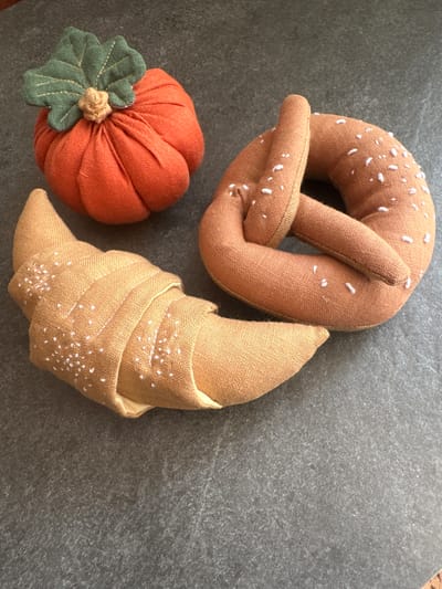 A fabric pumpkin with green leaves sits on a dark gray surface next to a fabric pretzel and a fabric croissant. The pretzel and croissant are light brown with white specks that resemble salt. The surface appears to be a stone countertop. The scene is brightly lit and the objects are in focus. The location in Chornomorske, Ukraine, is not discernible from the image.