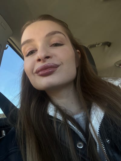 A young woman with long brown hair, glossy lips, and defined eyebrows is captured in an angled, close-up selfie. She wears a dark jacket with a white-lined zipper. The setting appears to be inside a car, evidenced by the vehicle's interior roof and a glimpse of the blue sky visible through the window. The lighting suggests it's daytime.