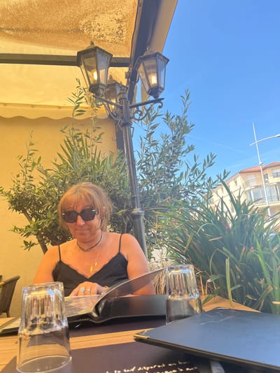 The image depicts an outdoor dining scene at a restaurant or cafe terrace in Saint-Just-Saint-Rambert, France, under a clear blue sky. A middle-aged woman is seated at a dark-colored table in the left half of the frame. She has shoulder-length, light brown hair with visible grey strands and wears dark sunglasses, a black spaghetti-strap top with lace trim, a black choker, and a gold necklace with a small pendant. Her left hand, adorned with multiple silver rings, rests on an open black menu or binder. Two inverted clear drinking glasses are visible on the table in the foreground.

Behind the woman, a light yellow building wall is partially obscured by lush green olive trees and other foliage. A prominent, ornate black metal street lamp with multiple lantern-style lights stands to the right of the woman, with one illuminated bulb visible. To the right of the table, a closed black menu or binder is present.

In the background, beyond the vegetation and lamp, multi-story buildings with light-colored facades, terracotta roofs, and balconies are visible under a clear blue sky. A white pole, possibly a streetlight or mast, extends into the sky on the far right. A faint aircraft contrail is discernible in the upper right sky. The scene is brightly lit by natural sunlight, indicating daytime.